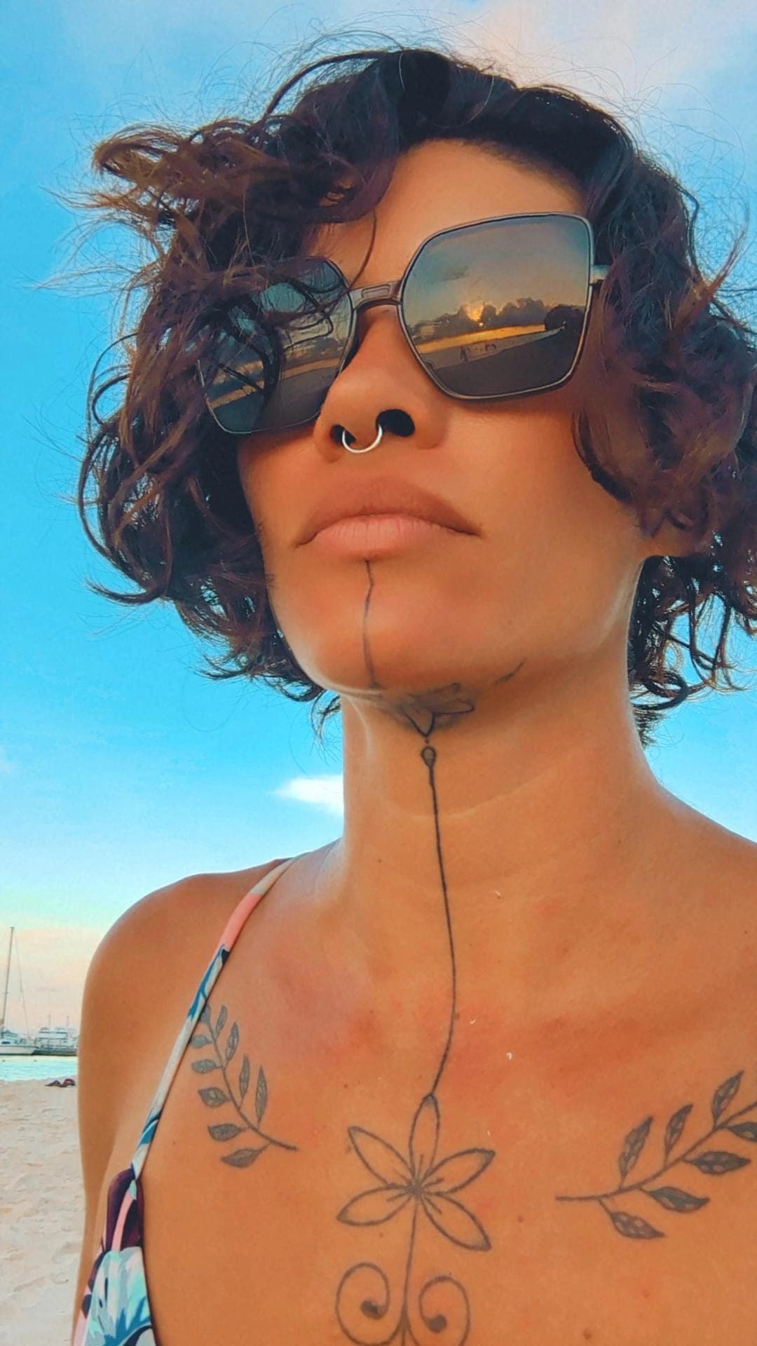 Woman with curly hair, sunglasses reflecting a sunset, and tattoos on her chin and chest.