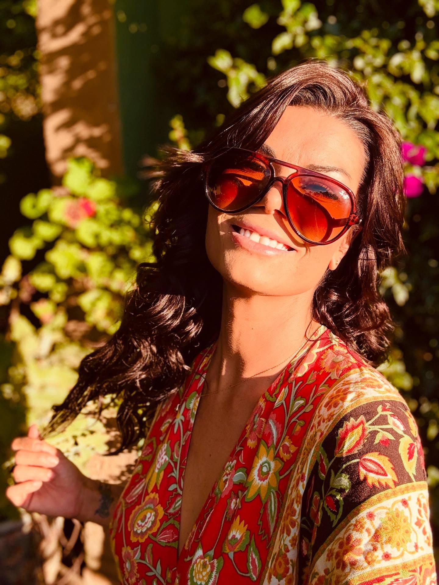Smiling woman with long dark wavy hair wearing oversized sunglasses and a colorful floral dress.
