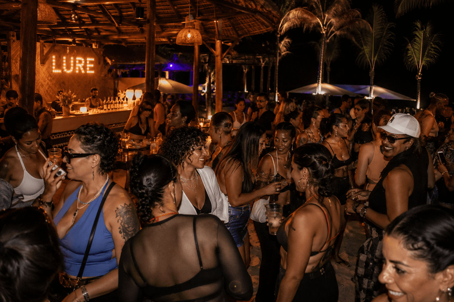Crowded outdoor tropical bar at night with people socializing under warm lights and palm trees.