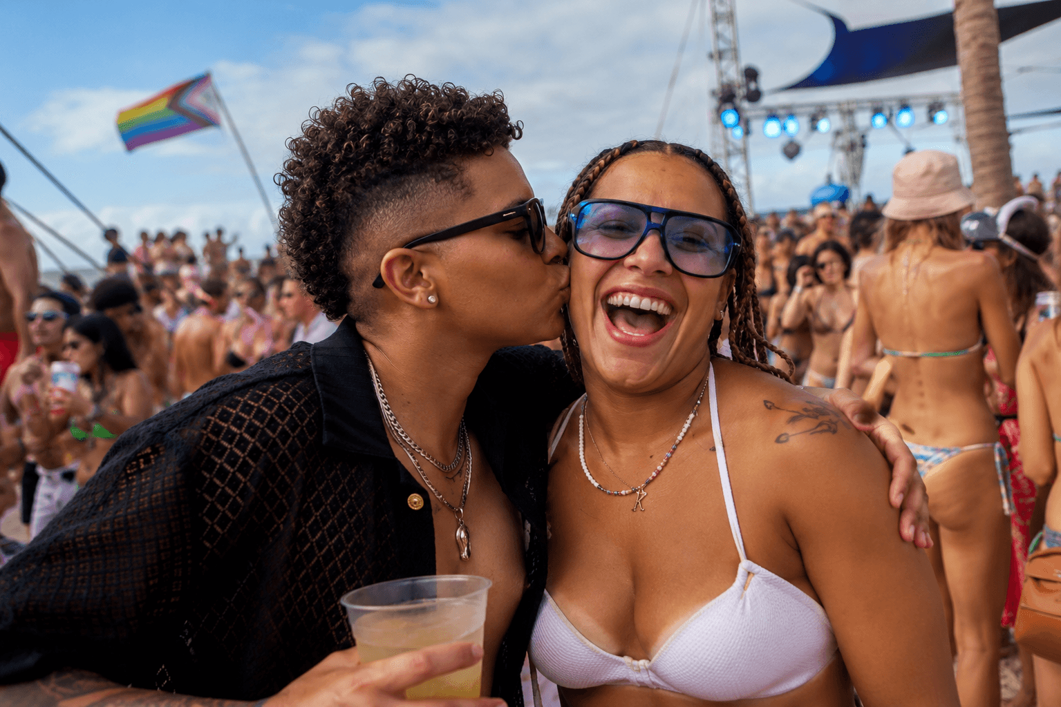 Person with curly hair kisses a smiling woman's cheek at a crowded outdoor pride festival.