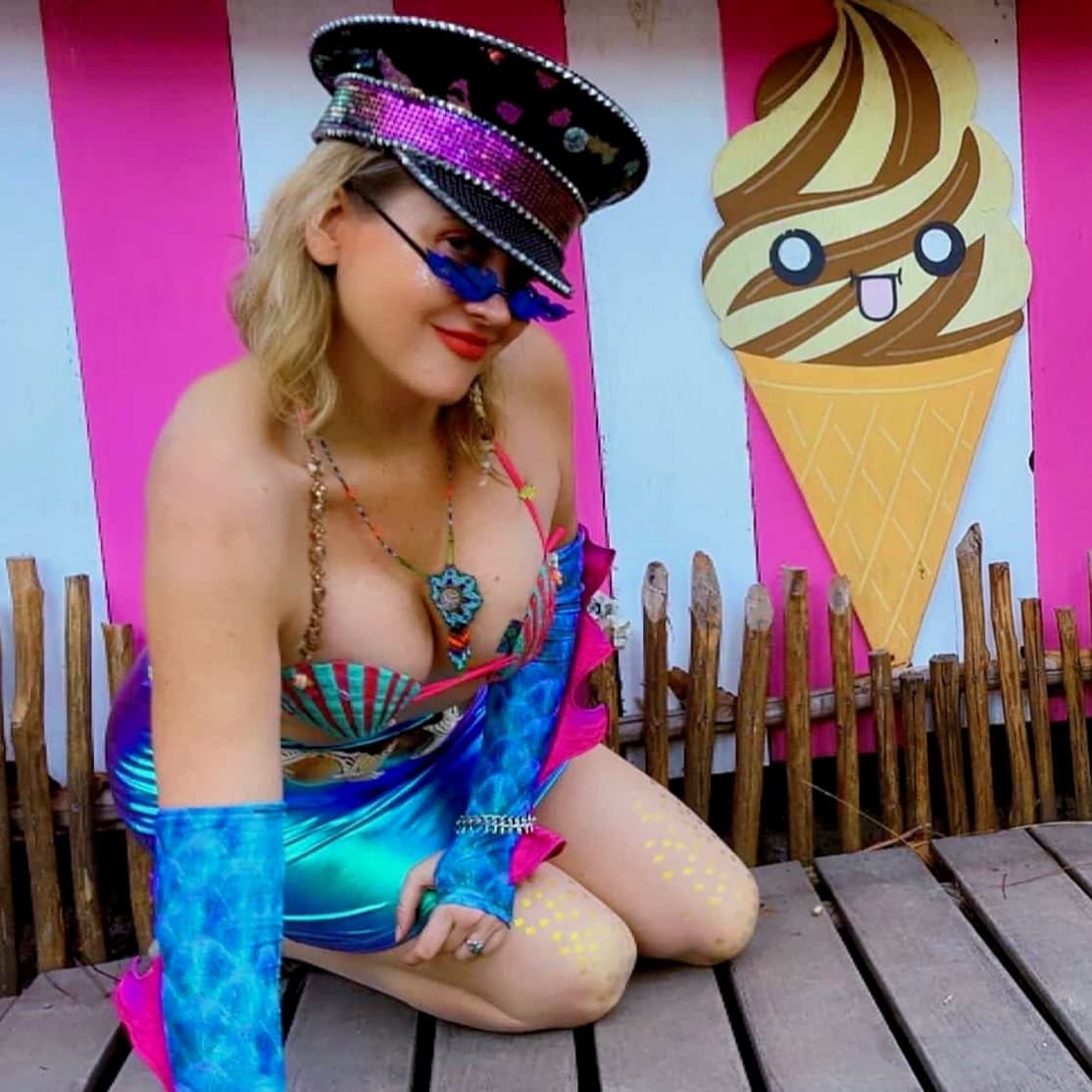 Woman in mermaid festival attire and sequined hat kneels beside a cartoon ice cream cutout.