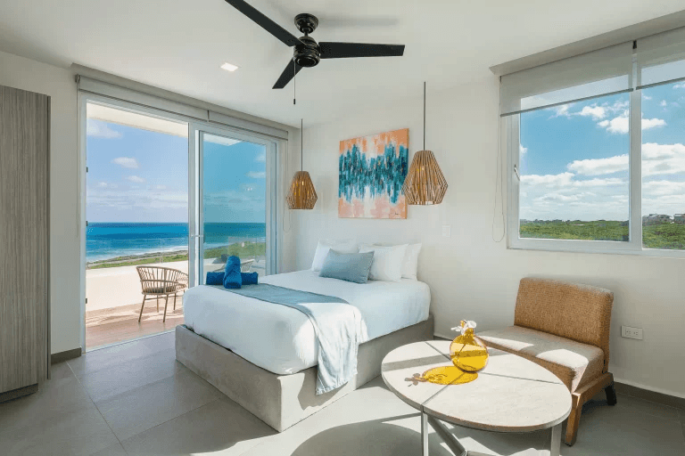 Modern bedroom with white bedding, blue accents, and a balcony overlooking the turquoise ocean.